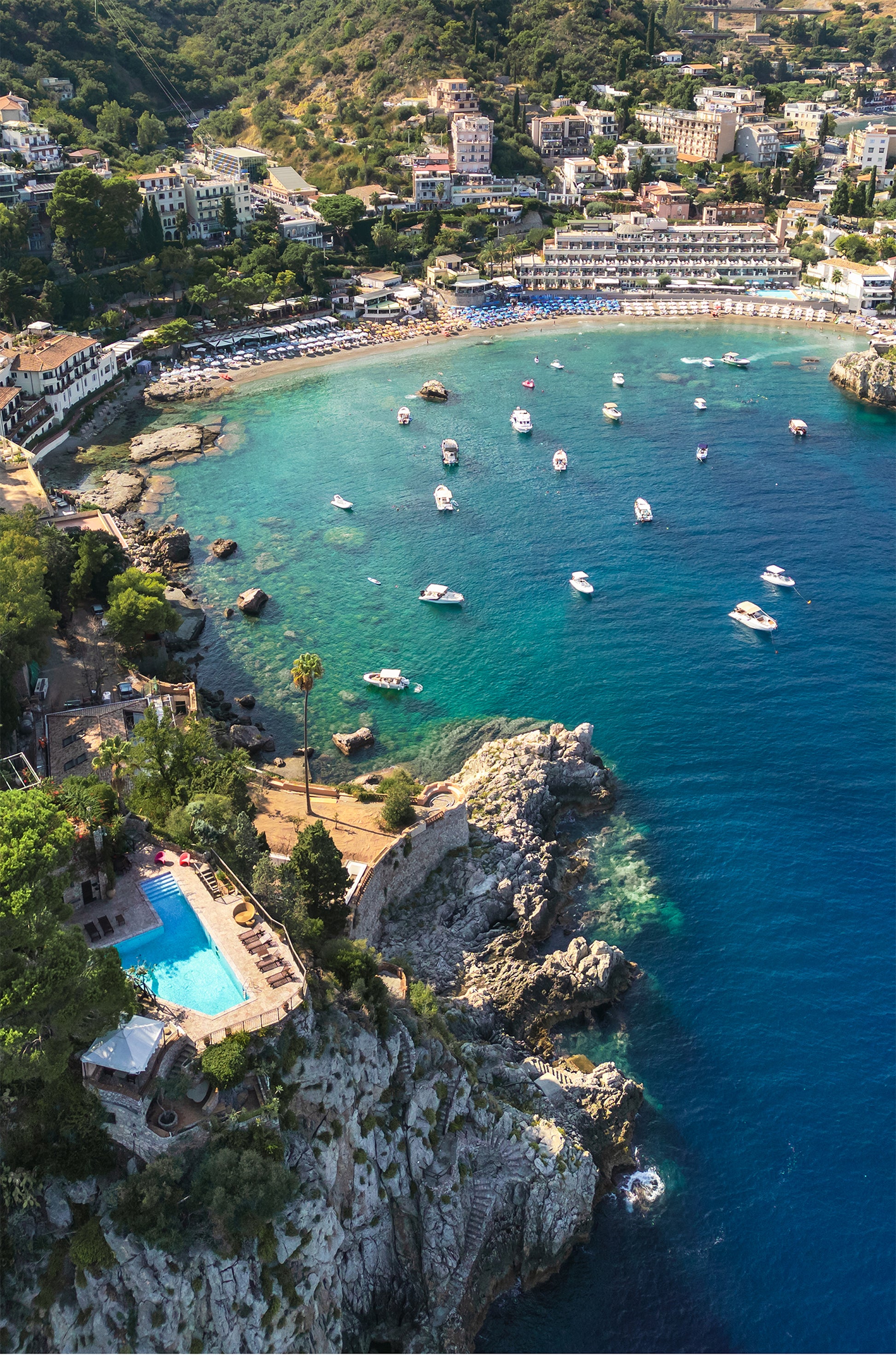 Aerial portrait view of Spiaggia di Mazzarò in Sicily, showcasing the curved coastline, turquoise bay, and surrounding cliffs.