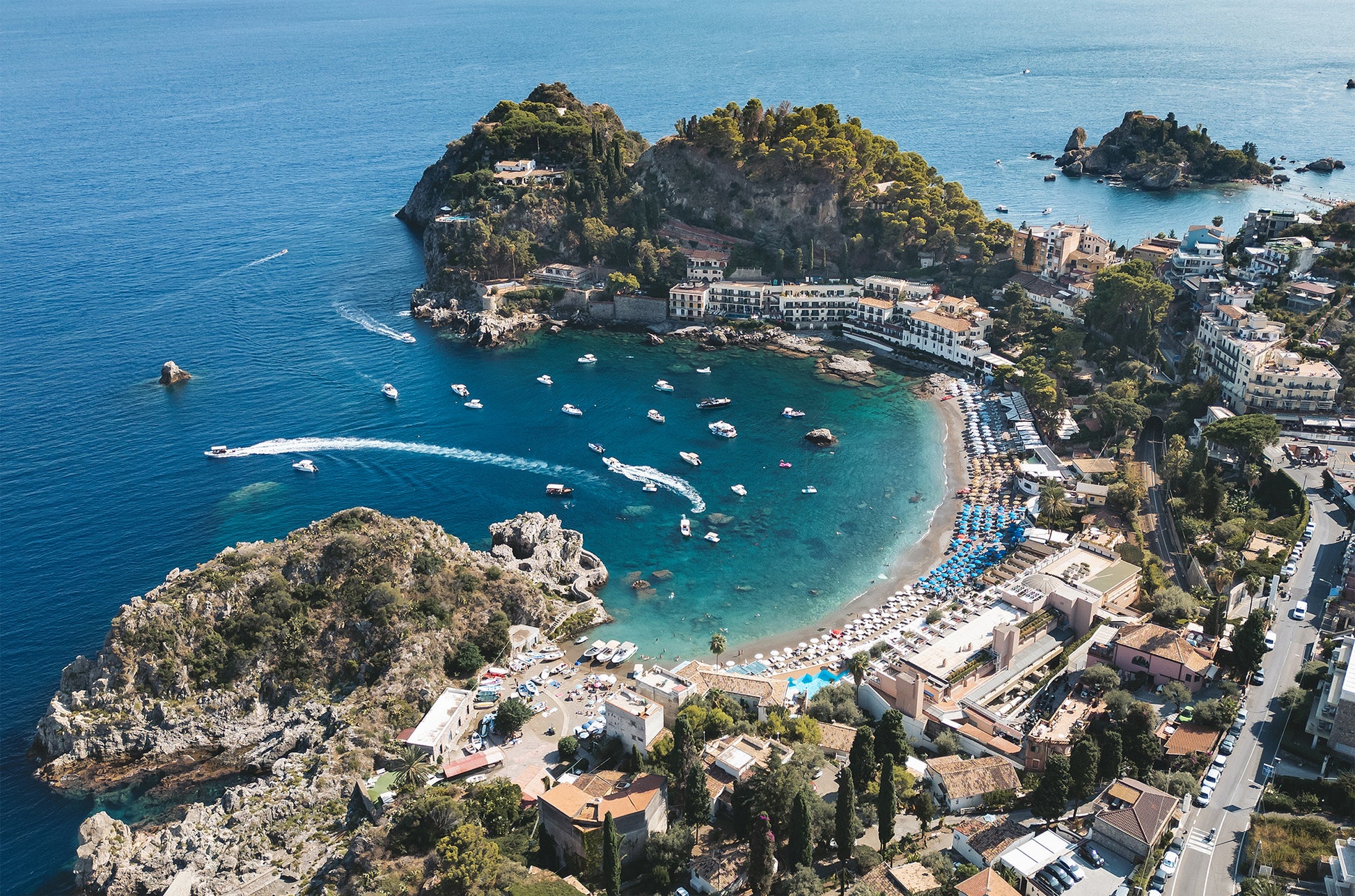 Landscape aerial photo of Spiaggia di Mazzarò in Sicily, with turquoise sea, sandy beach, and boats seen from an inland perspective.