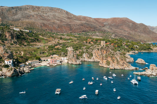 Drone photo of Scopello, Sicily showing the coastline, bay and boats docked on bright blue water.
