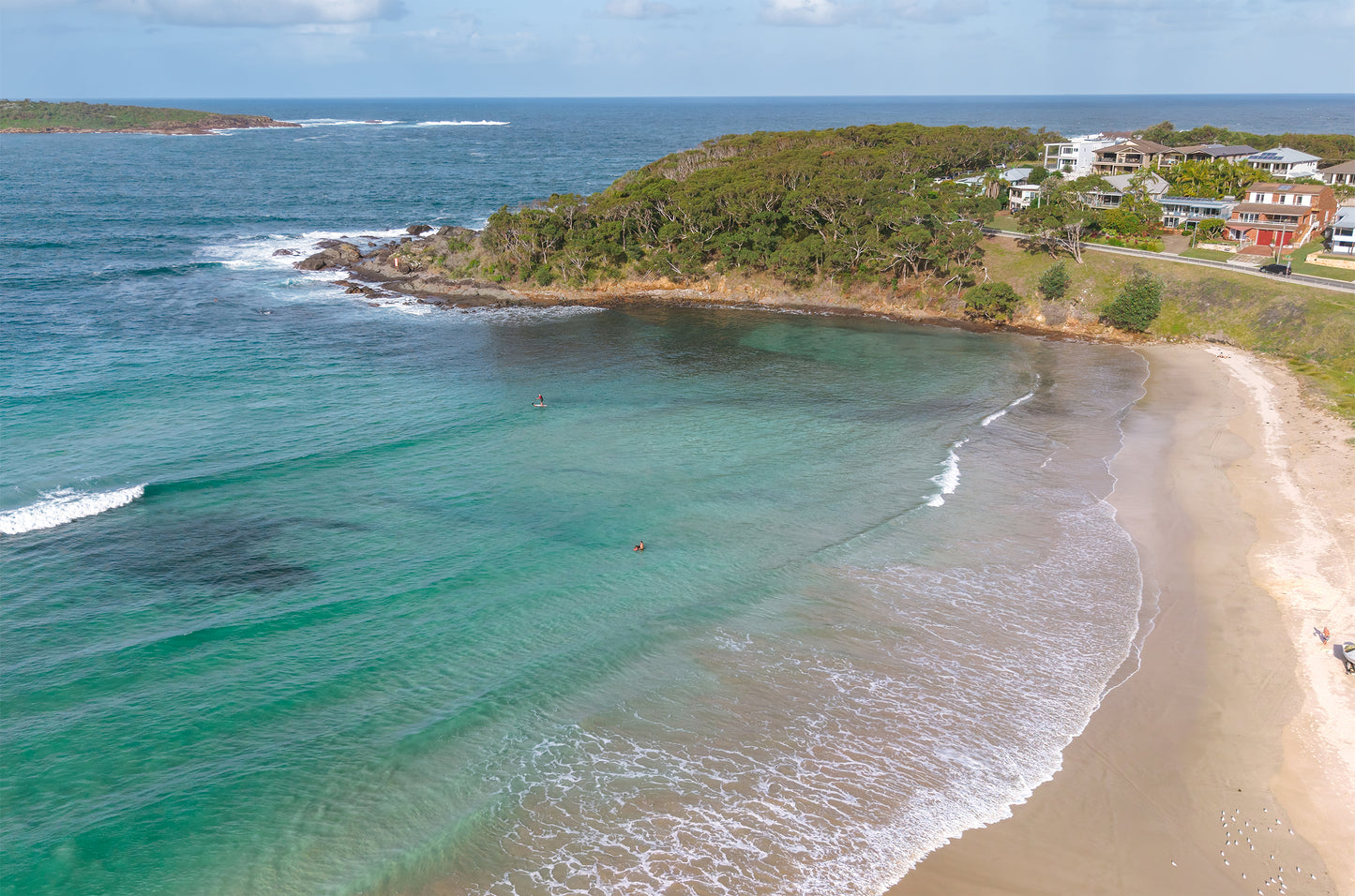 Aerial view of a sheltered cove at Fingal Bay near Port Stephens NSW, with emerald water, a paddleboarder and a bushland headland lined with beach houses.
