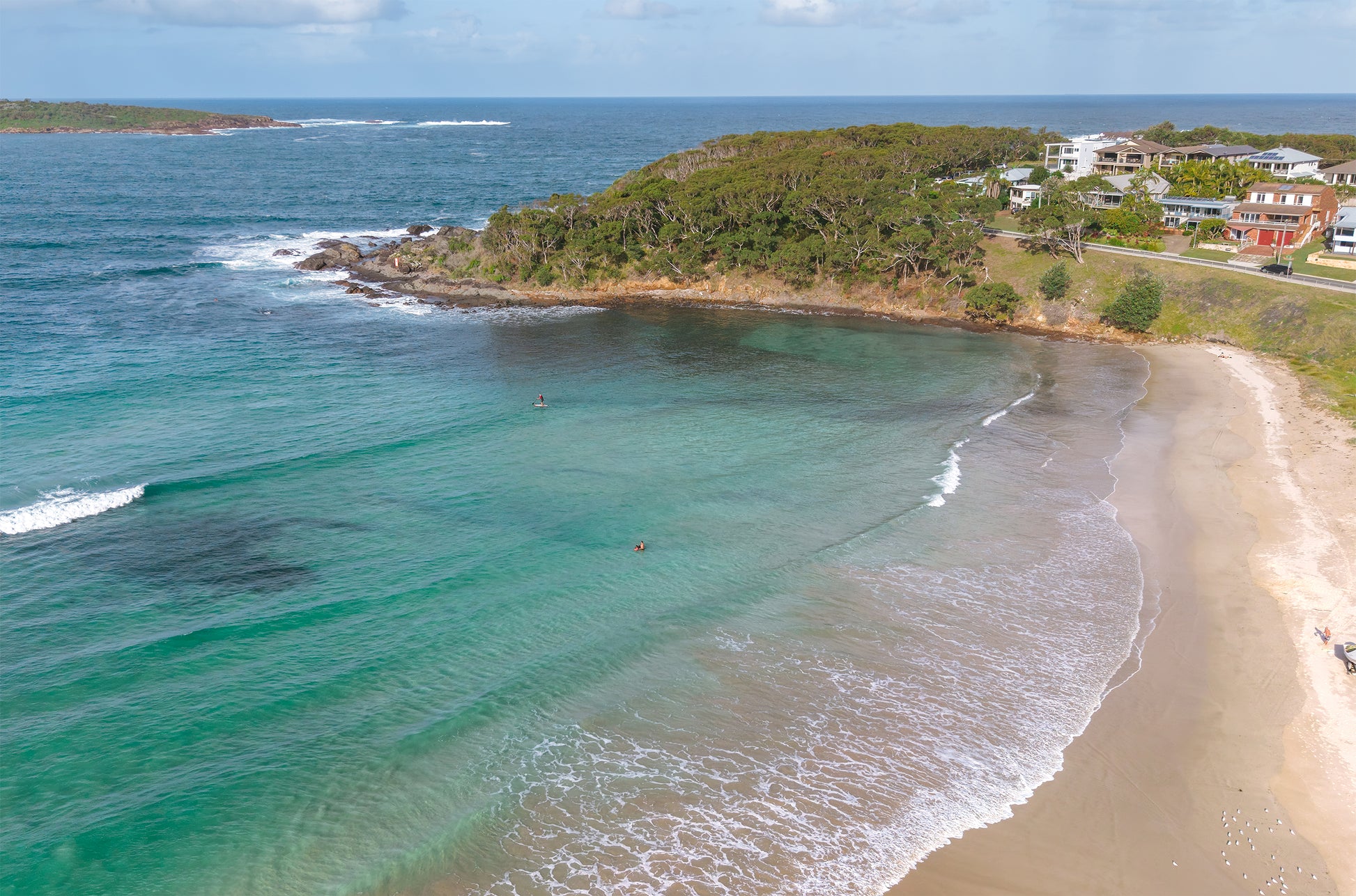 Aerial view of a sheltered cove at Fingal Bay near Port Stephens NSW, with emerald water, a paddleboarder and a bushland headland lined with beach houses.