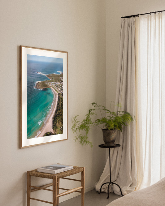 Fingal Bay cove portrait print in a natural oak frame, styled in a bedroom corner beside a wicker stool and fern.