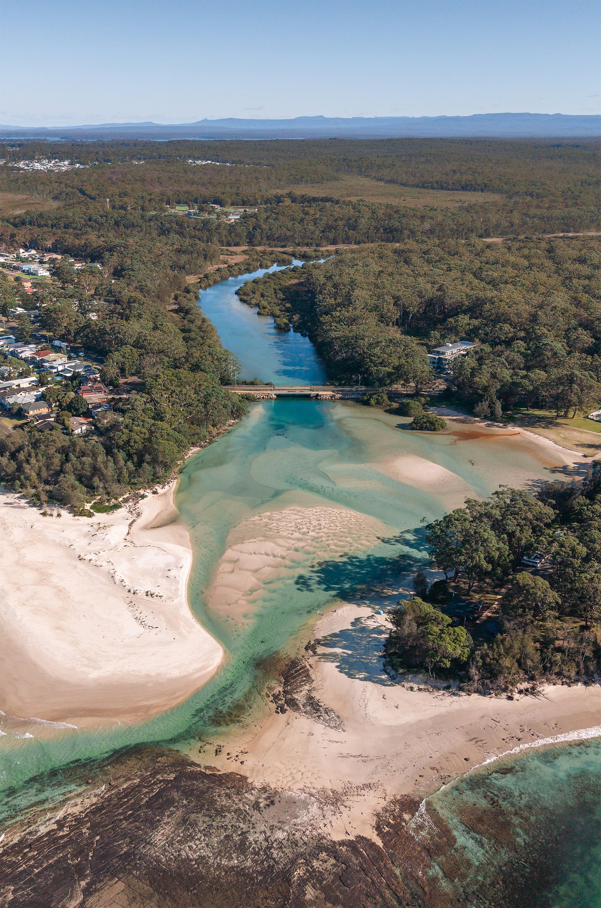 Aerial view of Moona Moona Creek near Huskisson NSW winding through bushland to Jervis Bay, with aqua channels, sandbars and mountains beyond.