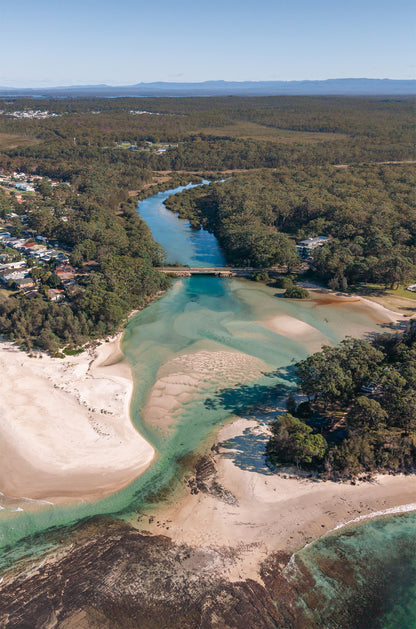 Aerial view of Moona Moona Creek near Huskisson NSW winding through bushland to Jervis Bay, with aqua channels, sandbars and mountains beyond.
