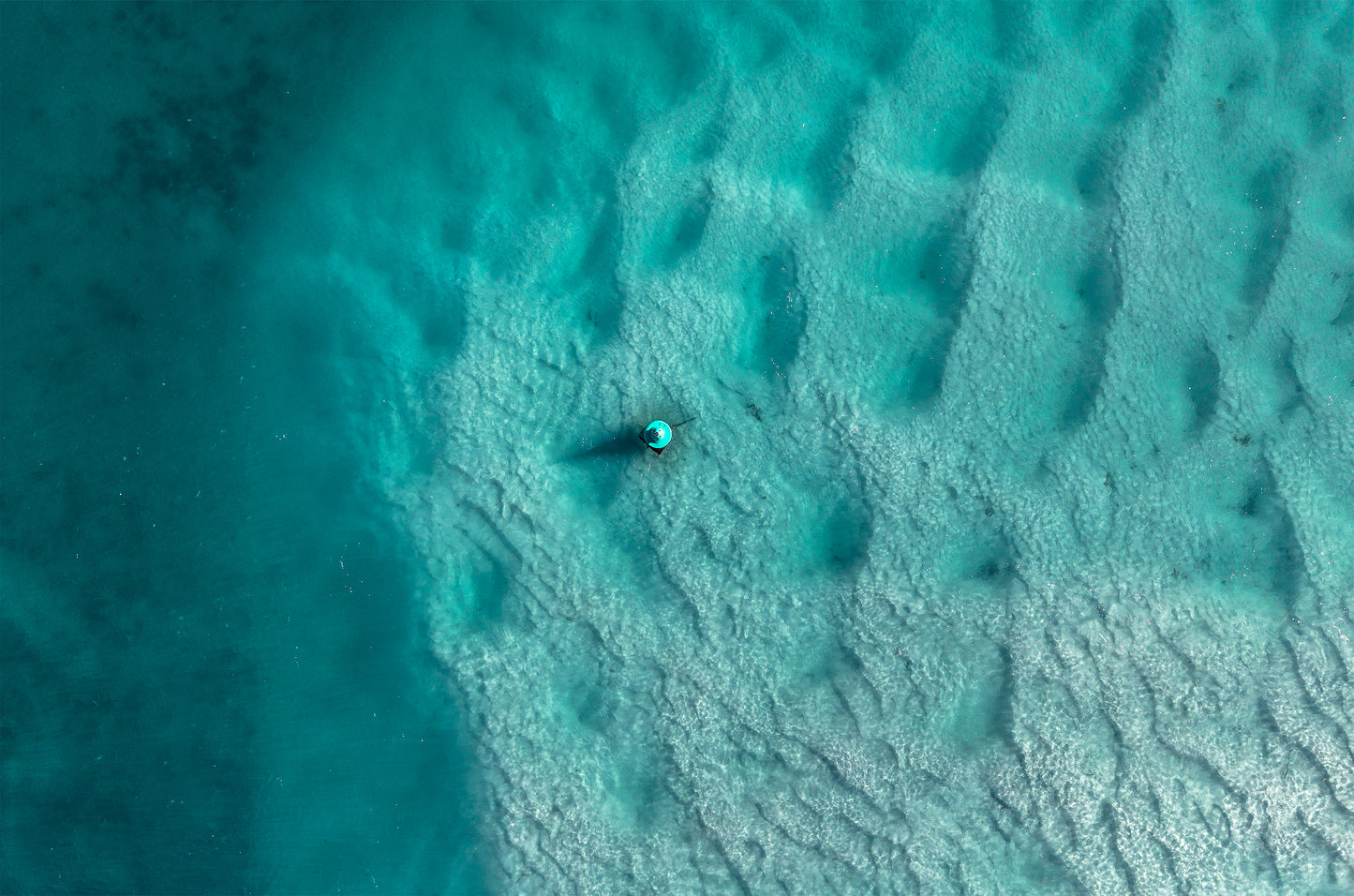 Top-down aerial of turquoise water over rippled sand in Currambene Creek, Huskisson NSW, with a single channel marker casting a shadow.