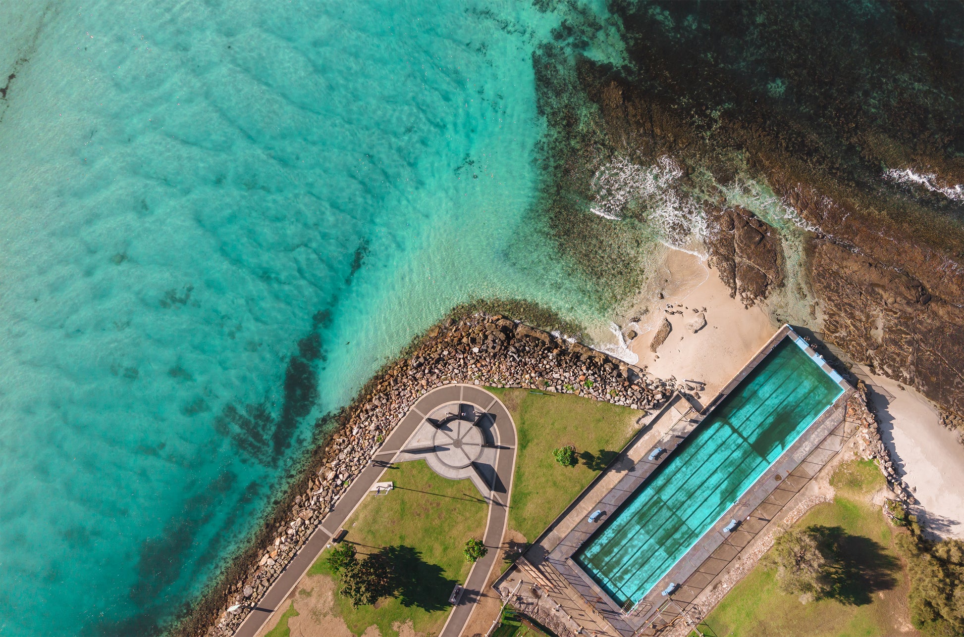 Top-down aerial of the Huskisson sea pool beside Jervis Bay NSW, with aqua pool water, rock wall, small beach and green lawns.