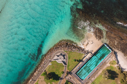 Top-down aerial of the Huskisson sea pool beside Jervis Bay NSW, with aqua pool water, rock wall, small beach and green lawns.