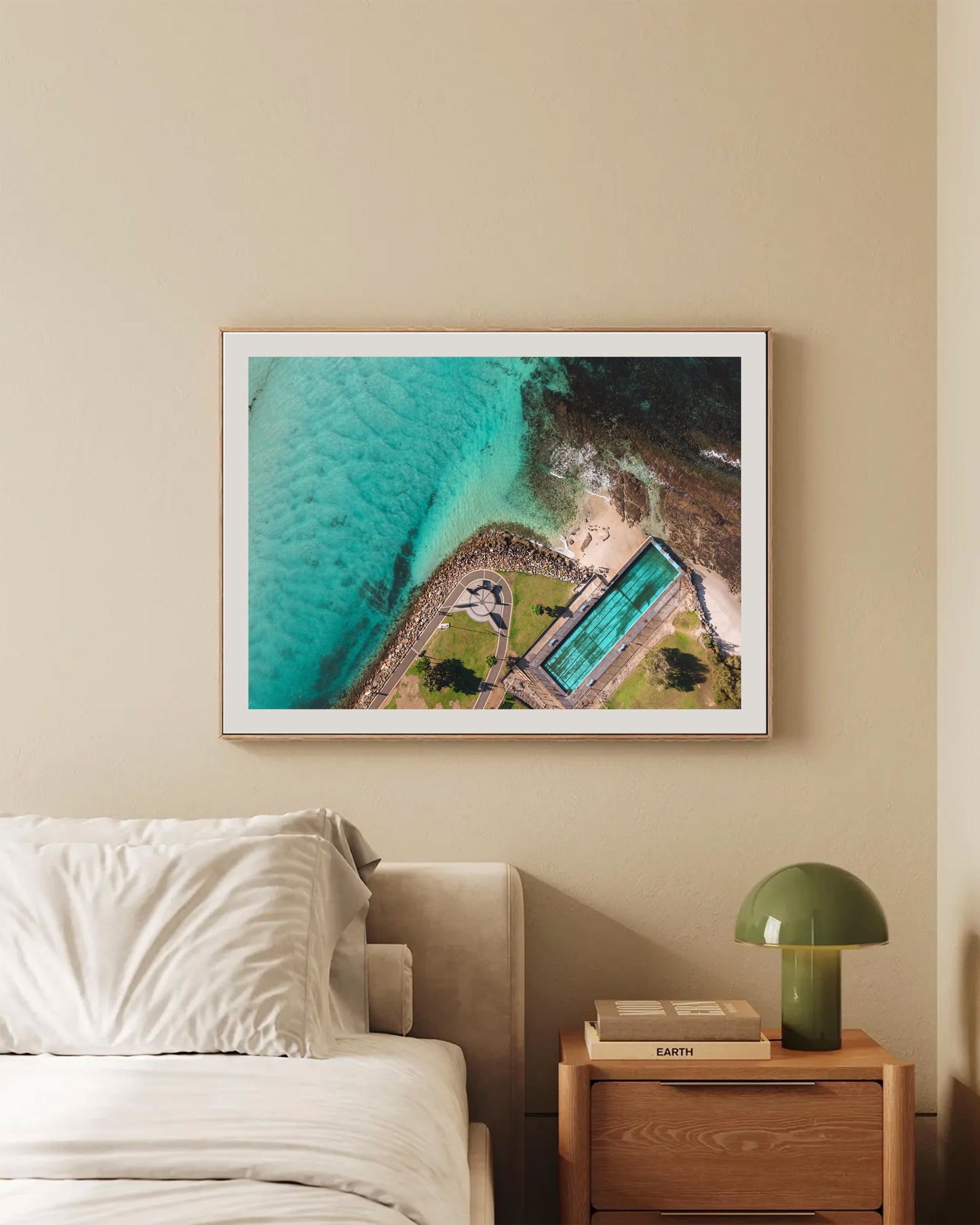 Huskisson Sea Pool print in a natural oak frame, styled above a bed in a warm neutral bedroom.