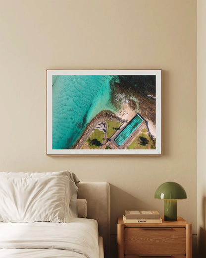 Huskisson Sea Pool print in a natural oak frame, styled above a bed in a warm neutral bedroom.