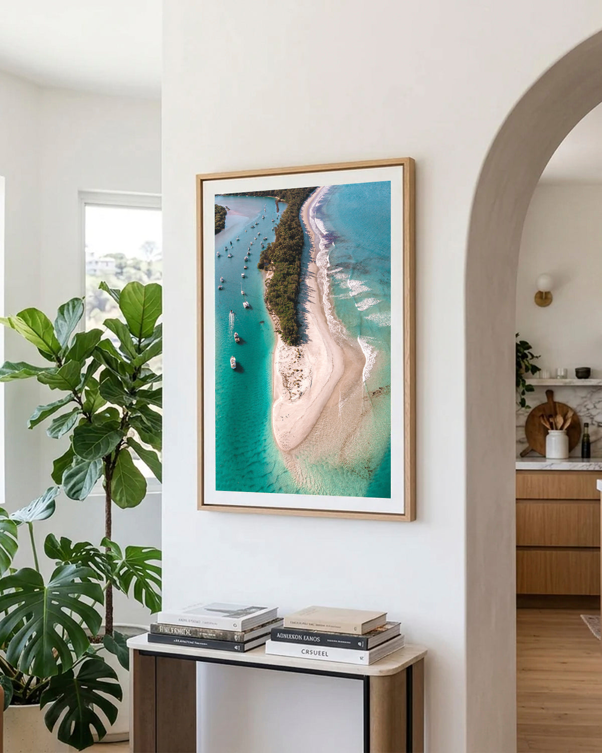 Myola Kite Beach print in a natural oak frame, styled in a hallway beside an arched doorway and indoor plants.