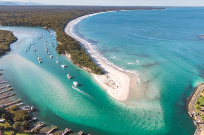 Wide aerial of Currambene Creek meeting Jervis Bay at Myola NSW, with moored yachts, a white sand spit and the long curve of the bay beyond.