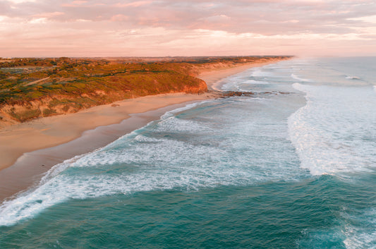 Sunset aerial of Kilcunda VIC with pink clouds above golden clifftops, teal surf rolling in and the Bass Coast fading into the distance.