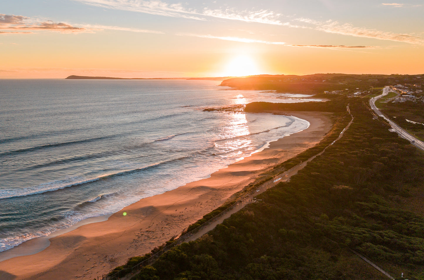 Golden hour aerial at Kilcunda VIC, with the sun setting over coastal cliffs and warm light across the surf beach and rolling waves.