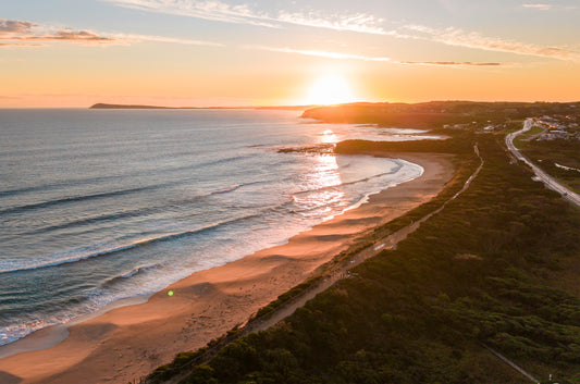 Golden hour aerial at Kilcunda VIC, with the sun setting over coastal cliffs and warm light across the surf beach and rolling waves.