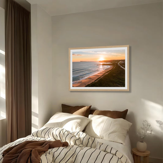 Kilcunda Golden Hour print in a natural oak frame, styled above a bed with striped linen in a soft evening-lit bedroom.