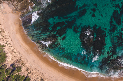 Top-down aerial of Kilcunda Beach VIC, where deep emerald water and dark reef meet a curve of golden sand laced with small waves.