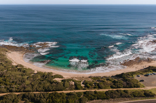 Aerial view of a horseshoe-shaped cove at Kilcunda VIC, with turquoise water over reef, golden sand and the coastal rail trail above the beach.