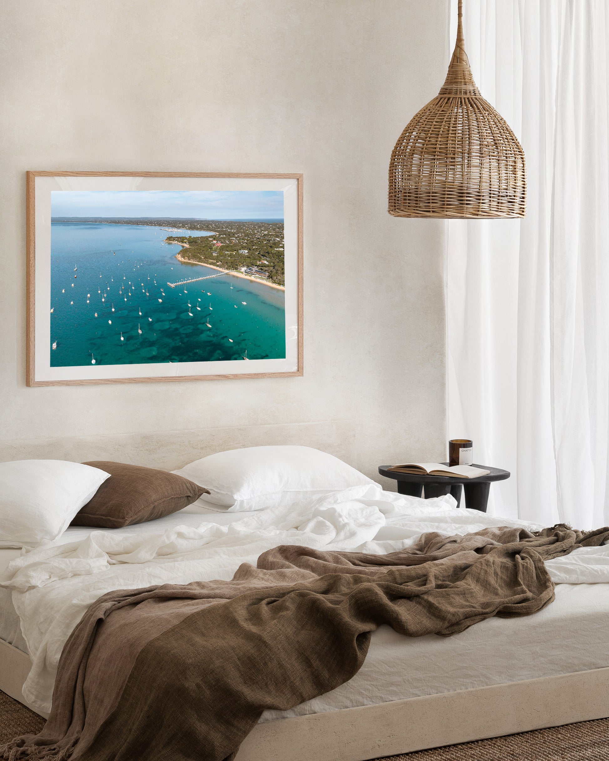 Sorrento Front Beach print in a natural oak frame, styled above a bed with brown linen cushions beneath a wicker pendant.