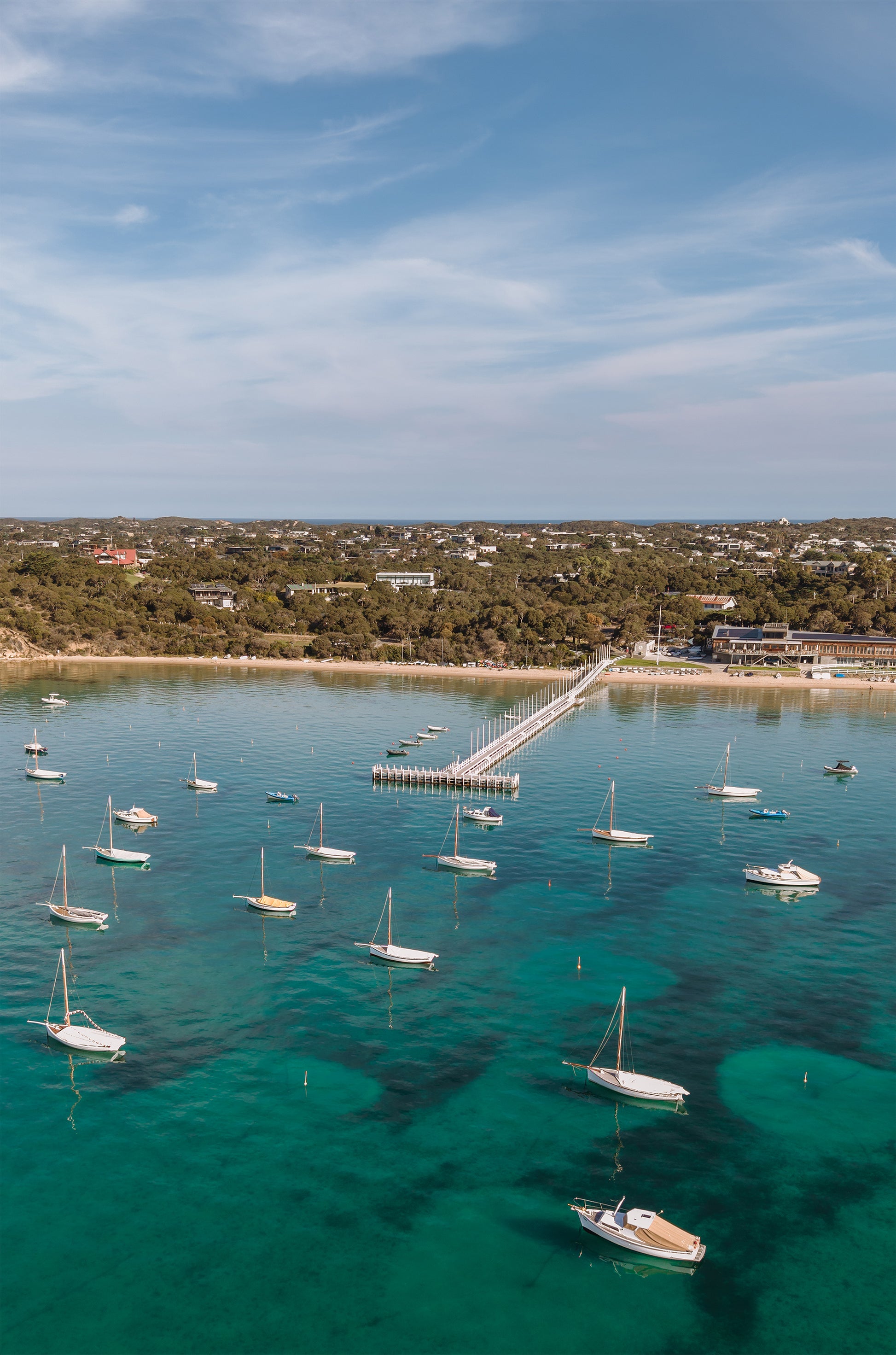 Portrait aerial of couta boats and yachts moored near the Sorrento Sailing Couta Boat Club jetty on teal Port Phillip Bay water.