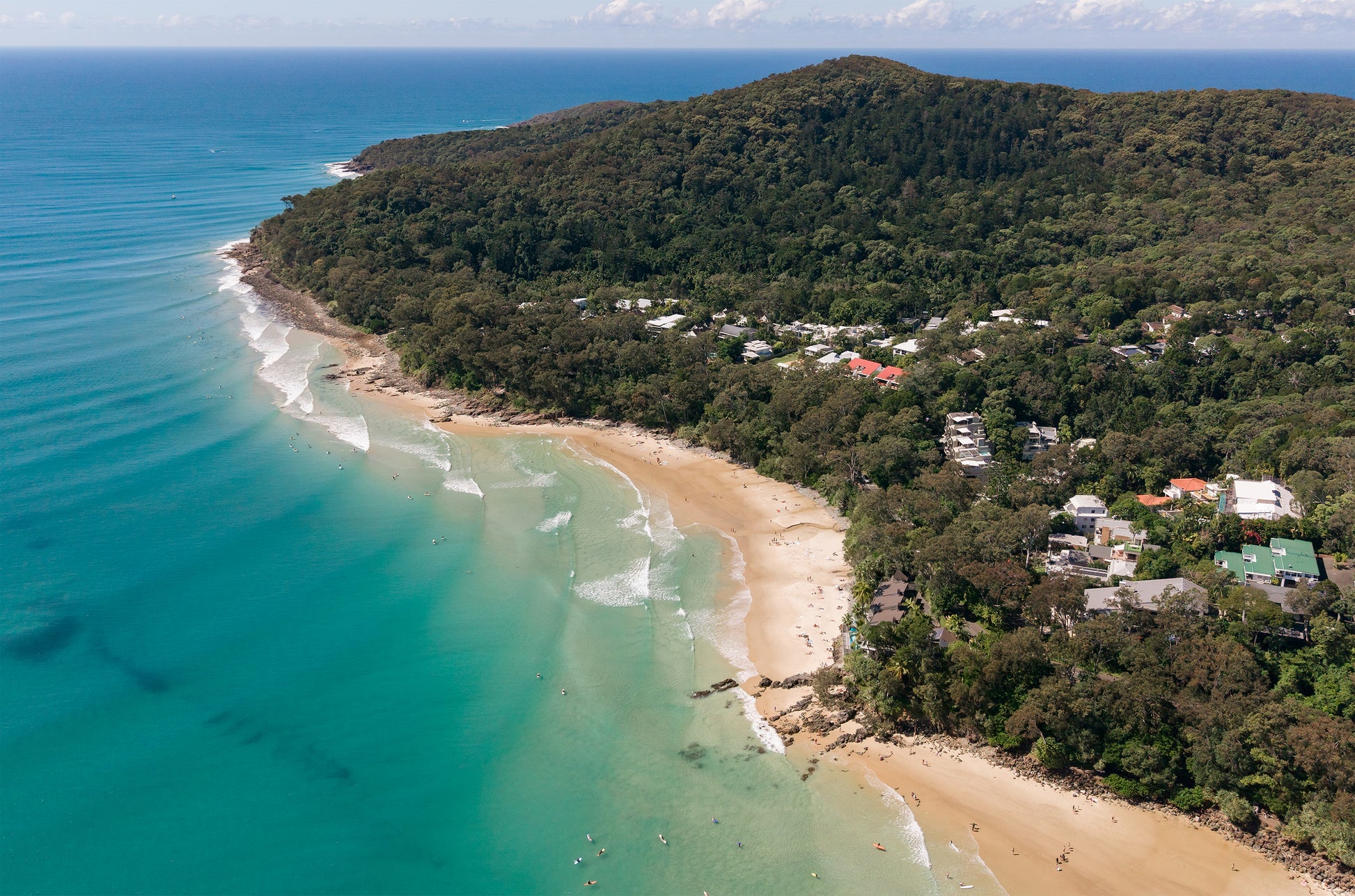 Aerial view of Little Cove and the Noosa National Park headland on the Sunshine Coast, with turquoise water, small waves and houses tucked into the rainforest.