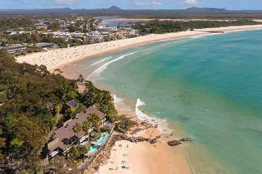 Aerial view over Little Cove to Noosa Main Beach and Laguna Bay on the Sunshine Coast, with crowds on the sand, surfers in turquoise water and the Noosa River beyond.