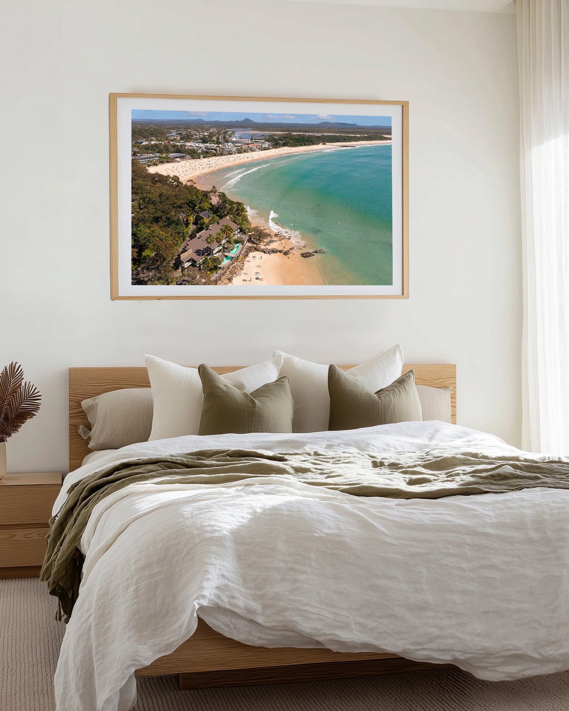 Noosa Main Beach and Laguna Bay aerial print in a natural oak frame, styled above a bed dressed in white and olive linen.