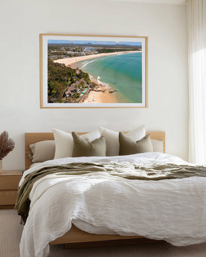 Noosa Main Beach and Laguna Bay aerial print in a natural oak frame, styled above a bed dressed in white and olive linen.