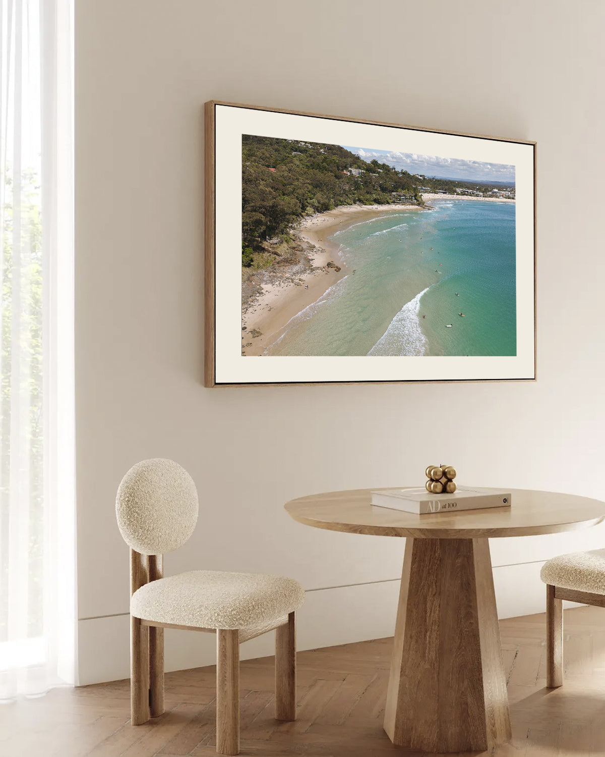 Little Cove Noosa aerial print framed and styled above a round timber dining table with bouclé chairs beside a sunlit window.