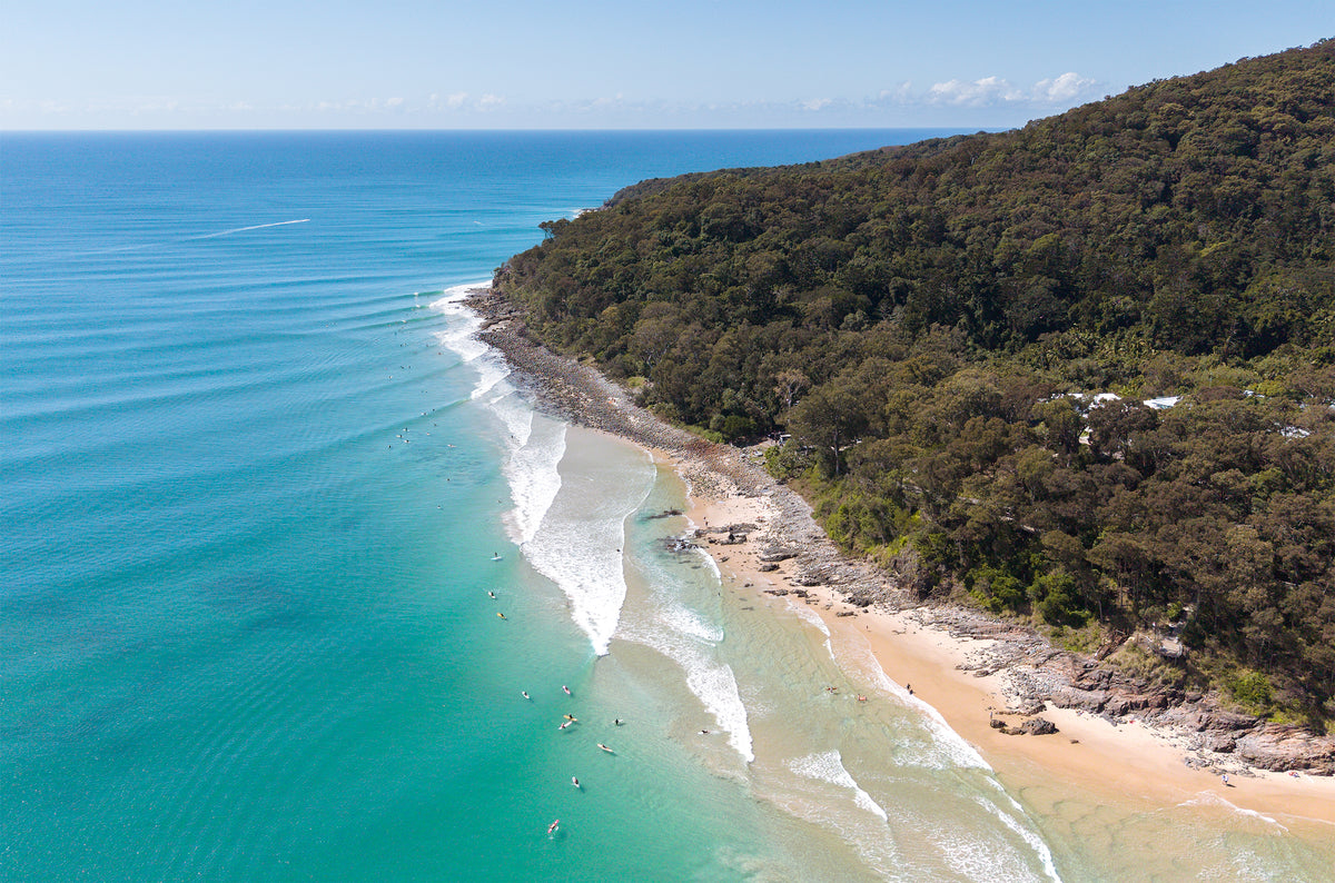Aerial view of the Noosa National Park headland on the Sunshine Coast, with rainforest, a rocky shoreline, surfers in the line-up and lines of swell over turquoise water.