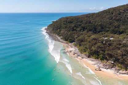 Aerial view of the Noosa National Park headland on the Sunshine Coast, with rainforest, a rocky shoreline, surfers in the line-up and lines of swell over turquoise water.