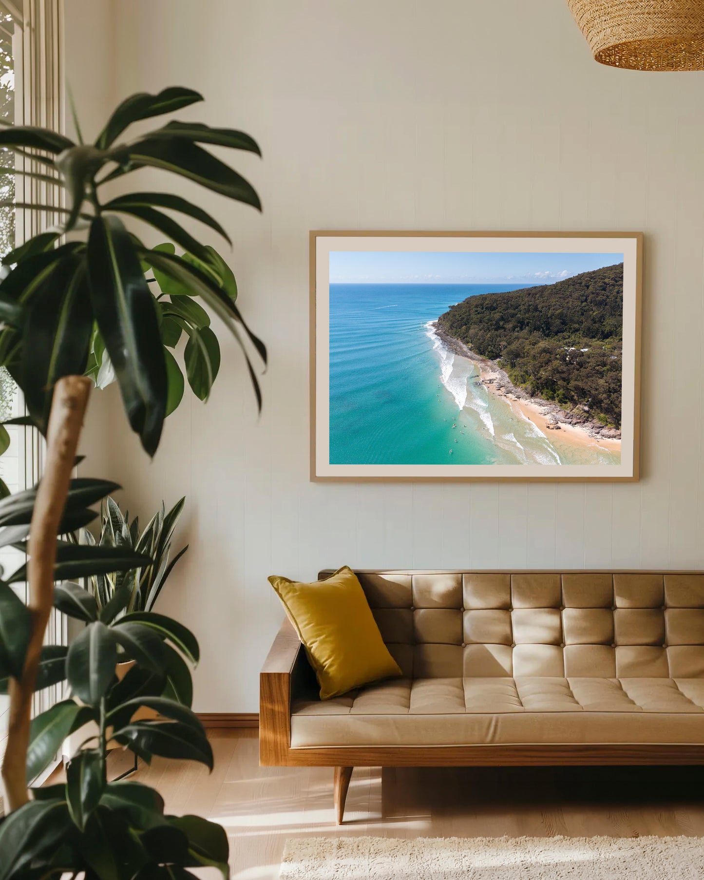 Noosa National Park Headland aerial print in a natural oak frame above a tan leather sofa with a mustard cushion and indoor plants.