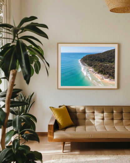 Noosa National Park Headland aerial print in a natural oak frame above a tan leather sofa with a mustard cushion and indoor plants.