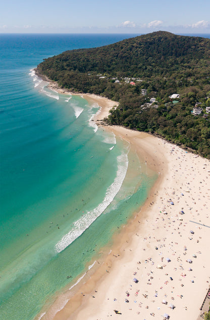 Portrait aerial view of Noosa Main Beach on the Sunshine Coast, with crowds of beachgoers on white sand, turquoise waves and the Noosa National Park headland beyond.
